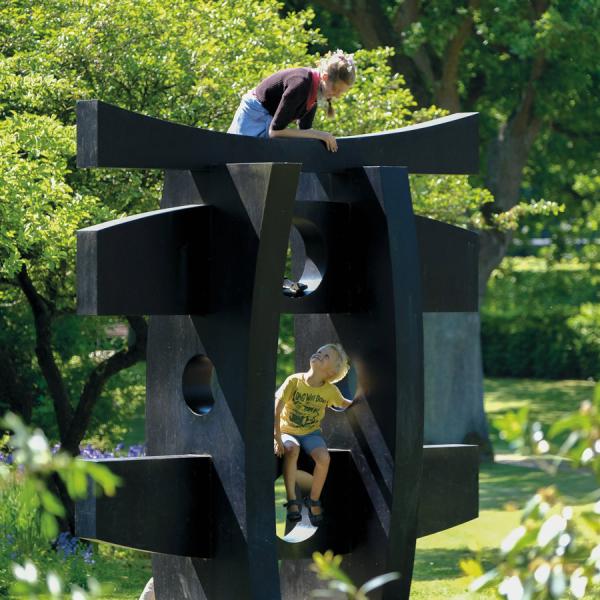 Zwei Kinder spielen auf der großen schwarzen Skulptur „Pagode“ von Hans August Andersen in Geografisk Have. Die Skulptur ist von üppiger Natur und grünen Bäumen umgeben. 